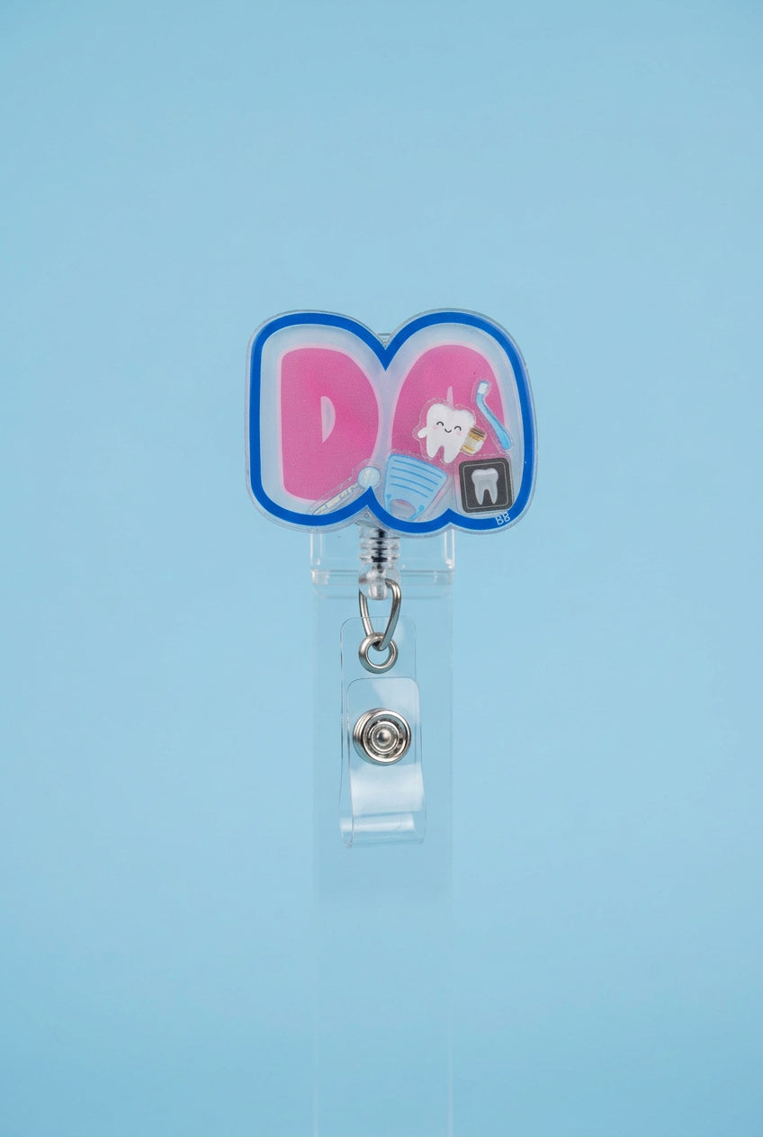 DA dental assistant shaker badge reel with pink lettering, a blue border, and miniature tooth, toothbrush, dental mirror, mask, and dental X-ray.