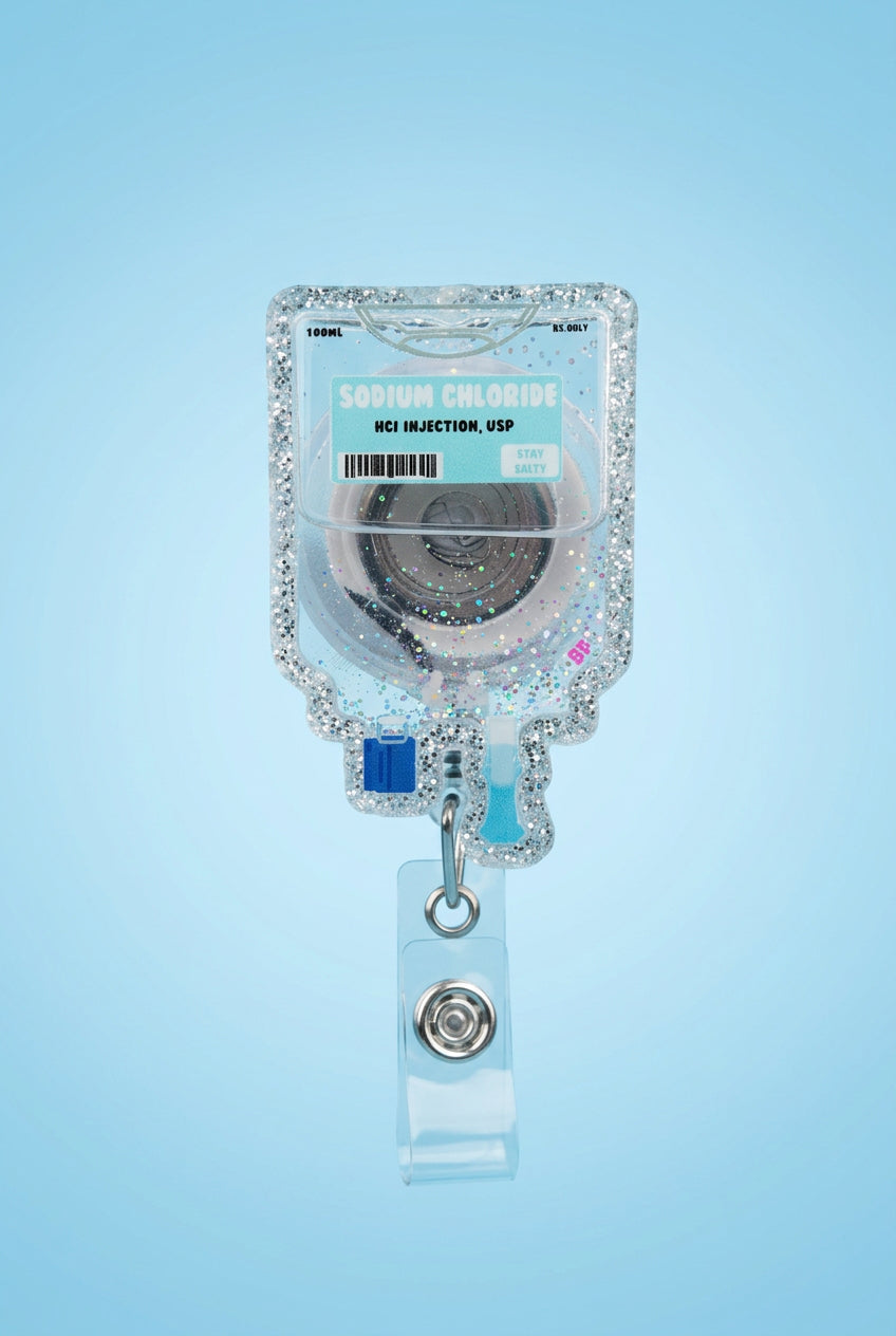 A hand wearing a pink glove holds a clear IV bag charm with pink glitter, a turquoise label reading Sodium Chloride, and a clear retractable badge clip on a gray wooden surface with a blurred outdoor background.