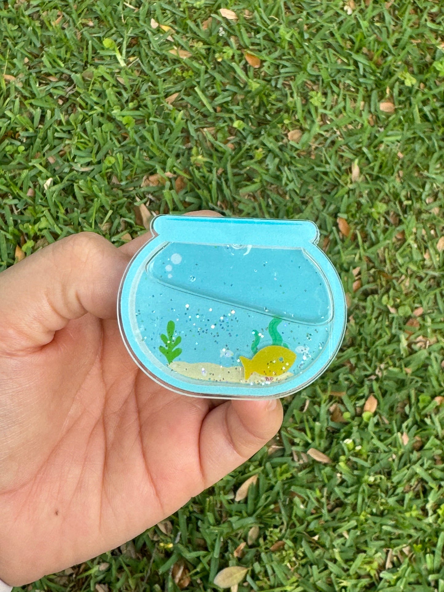 Blue fishbowl shaker badge reel featuring a yellow fish, aquatic plants, sandy bottom, and glitter accents