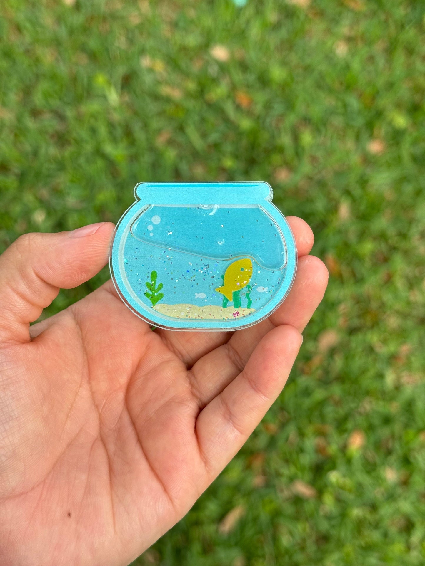 Blue fishbowl shaker badge reel featuring a yellow fish, aquatic plants, sandy bottom, and glitter accents