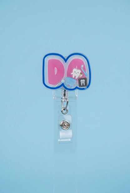 DA dental assistant shaker badge reel with pink lettering, a blue border, and miniature tooth, toothbrush, dental mirror, mask, and dental X-ray.