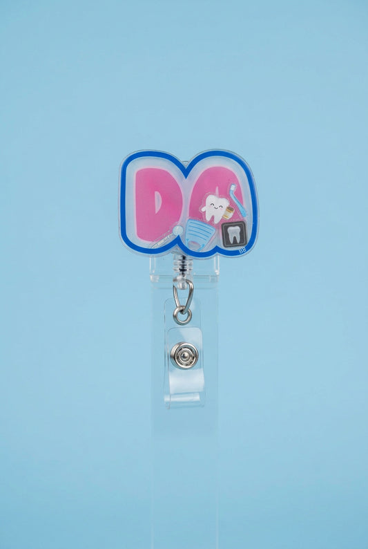 DA dental assistant shaker badge reel with pink lettering, a blue border, and miniature tooth, toothbrush, dental mirror, mask, and dental X-ray.