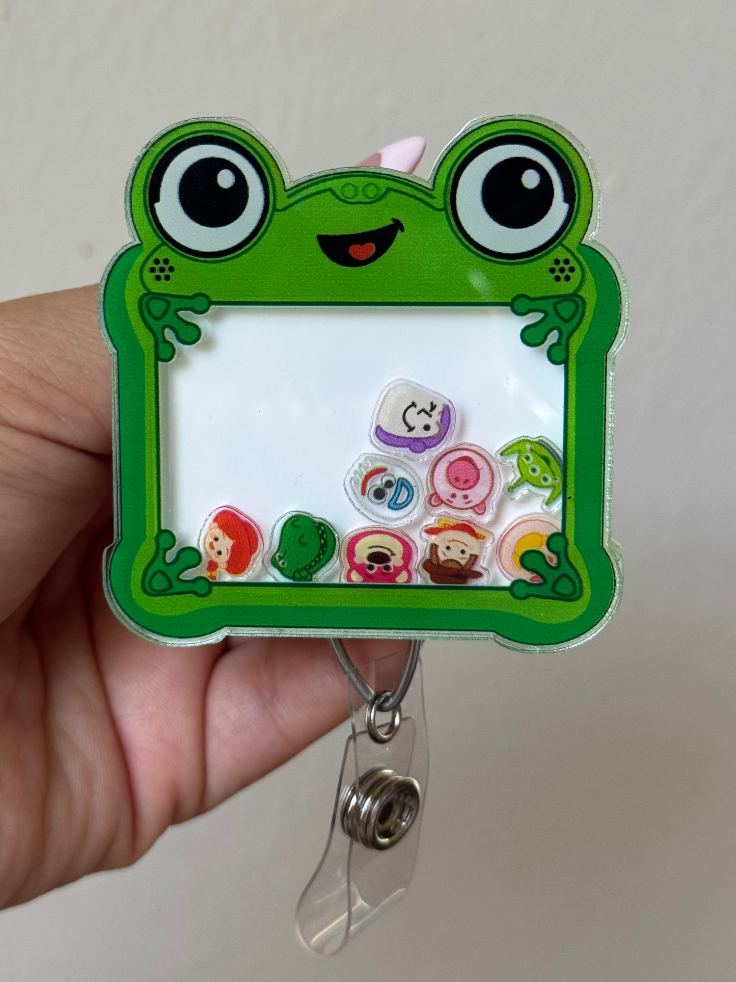 A hand holding a green frog-themed acrylic badge reel. The badge reel features a cartoon frog face, large round eyes, and a white writing pad with colorful stickers inside.
