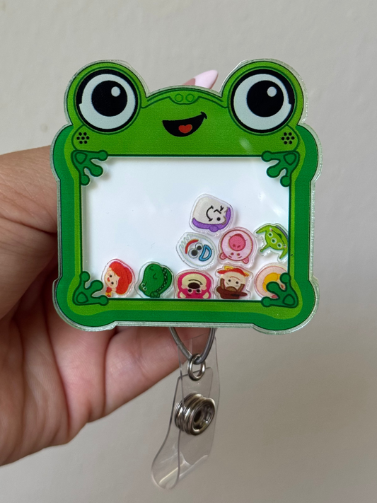 A hand holding a green frog-themed acrylic badge reel. The badge reel features a cartoon frog face, large round eyes, and a white writing pad with colorful stickers inside.