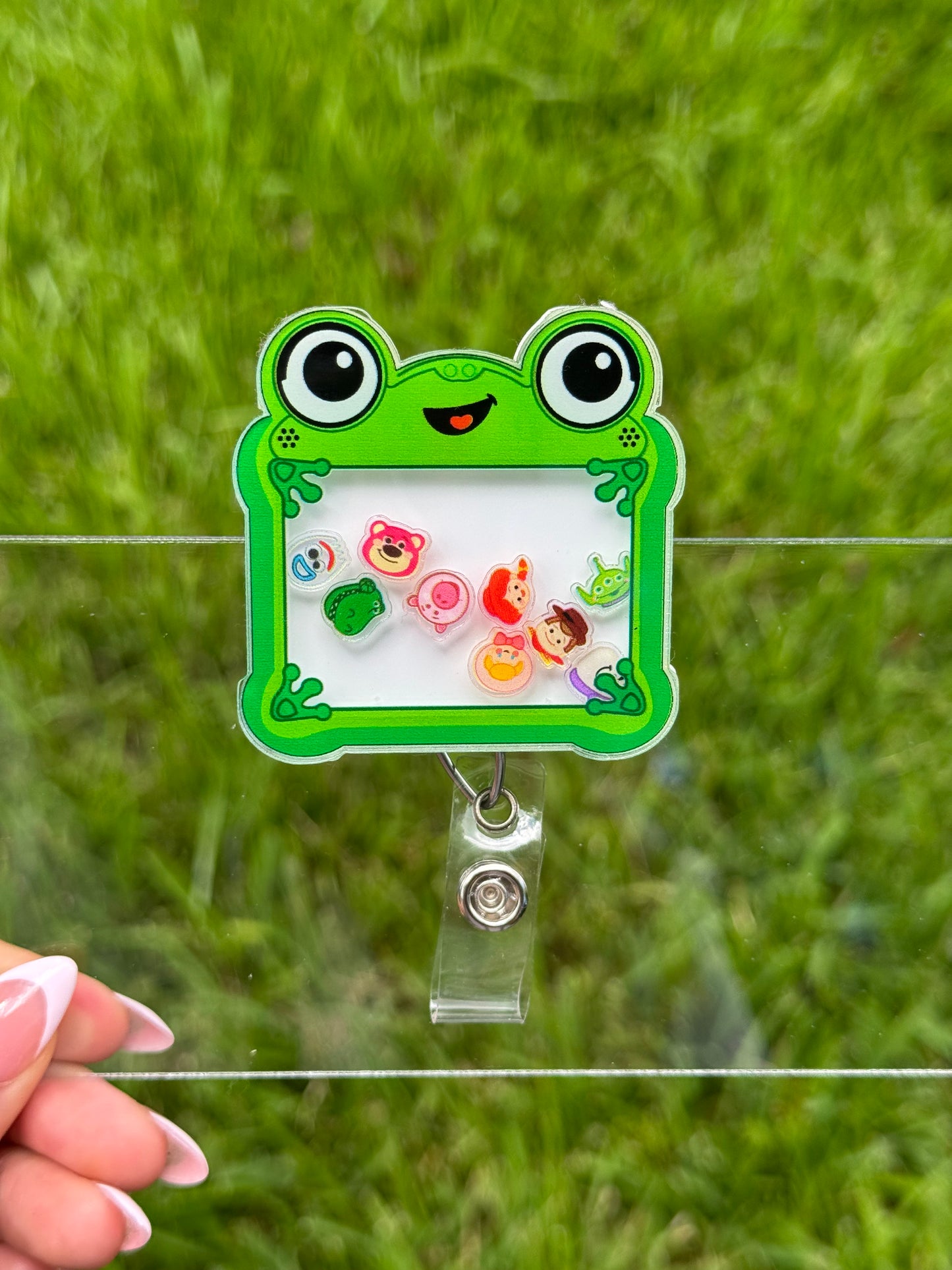 A green frog-shaped acrylic badge holder with a clear clip, held by a hand in front of a blurred green grass background.