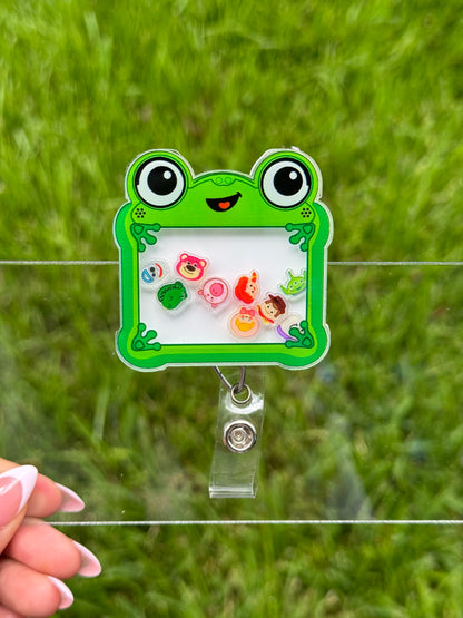 A green frog-shaped acrylic badge holder with a clear clip, held by a hand in front of a blurred green grass background.