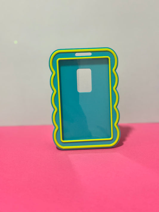 A translucent teal id holder with a bright yellow scalloped border, standing upright on a pink surface against a light gray background.