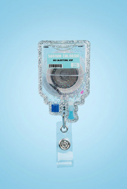 A hand wearing a pink glove holds a clear IV bag charm with pink glitter, a turquoise label reading Sodium Chloride, and a clear retractable badge clip on a gray wooden surface with a blurred outdoor background.