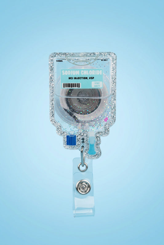 A hand wearing a pink glove holds a clear IV bag charm with pink glitter, a turquoise label reading Sodium Chloride, and a clear retractable badge clip on a gray wooden surface with a blurred outdoor background.