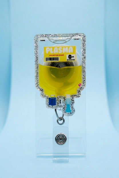 A novelty IV bag keychain or ID holder shaped like a glittery 'Plasma' bag filled with yellow liquid, held by a hand wearing a pink glove on a wooden deck.