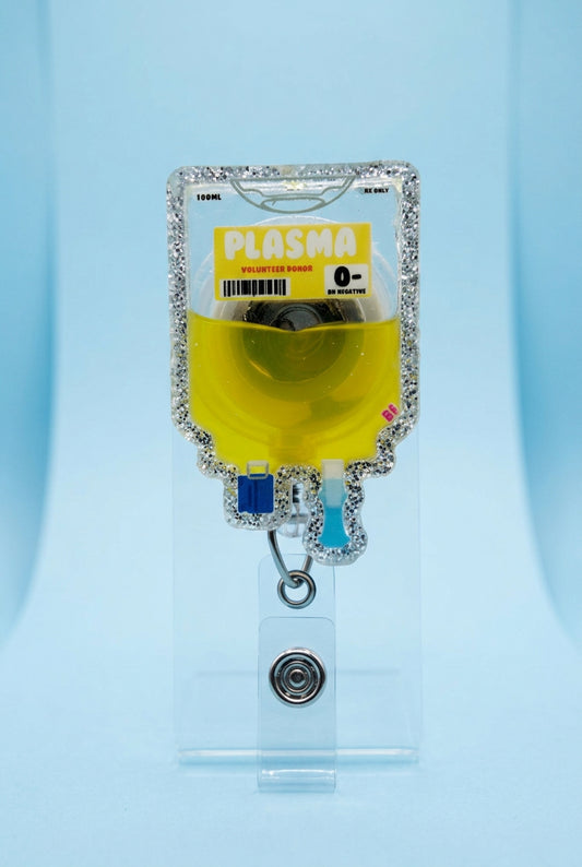 A novelty IV bag keychain or ID holder shaped like a glittery 'Plasma' bag filled with yellow liquid, held by a hand wearing a pink glove on a wooden deck.